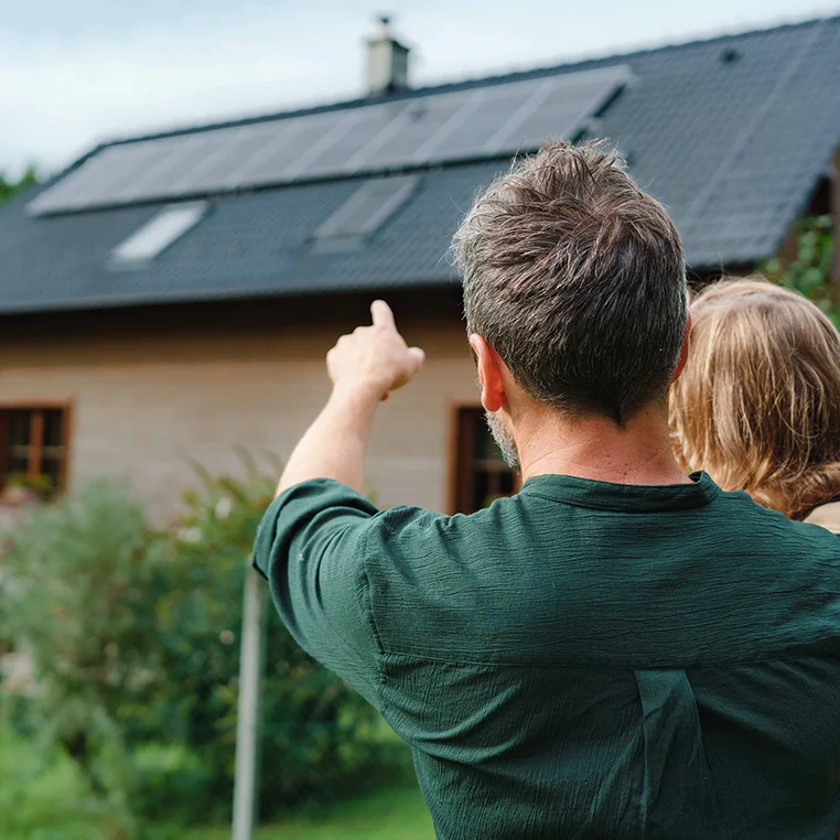 Mann mit Tochter im Arm deutet auf Photovoltaikanlage auf seinem Hausdach